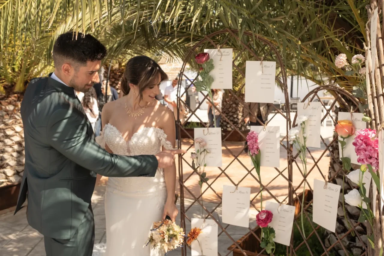 Pareja de recién casados observando un seating plan adornado con flores en una celebración al aire libre