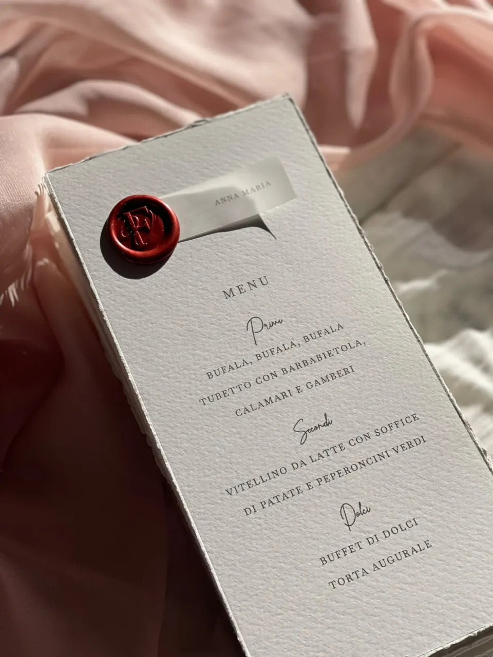 Minuta de boda sobre papel texturizado, con lacre granate y fondo de tela rosa
