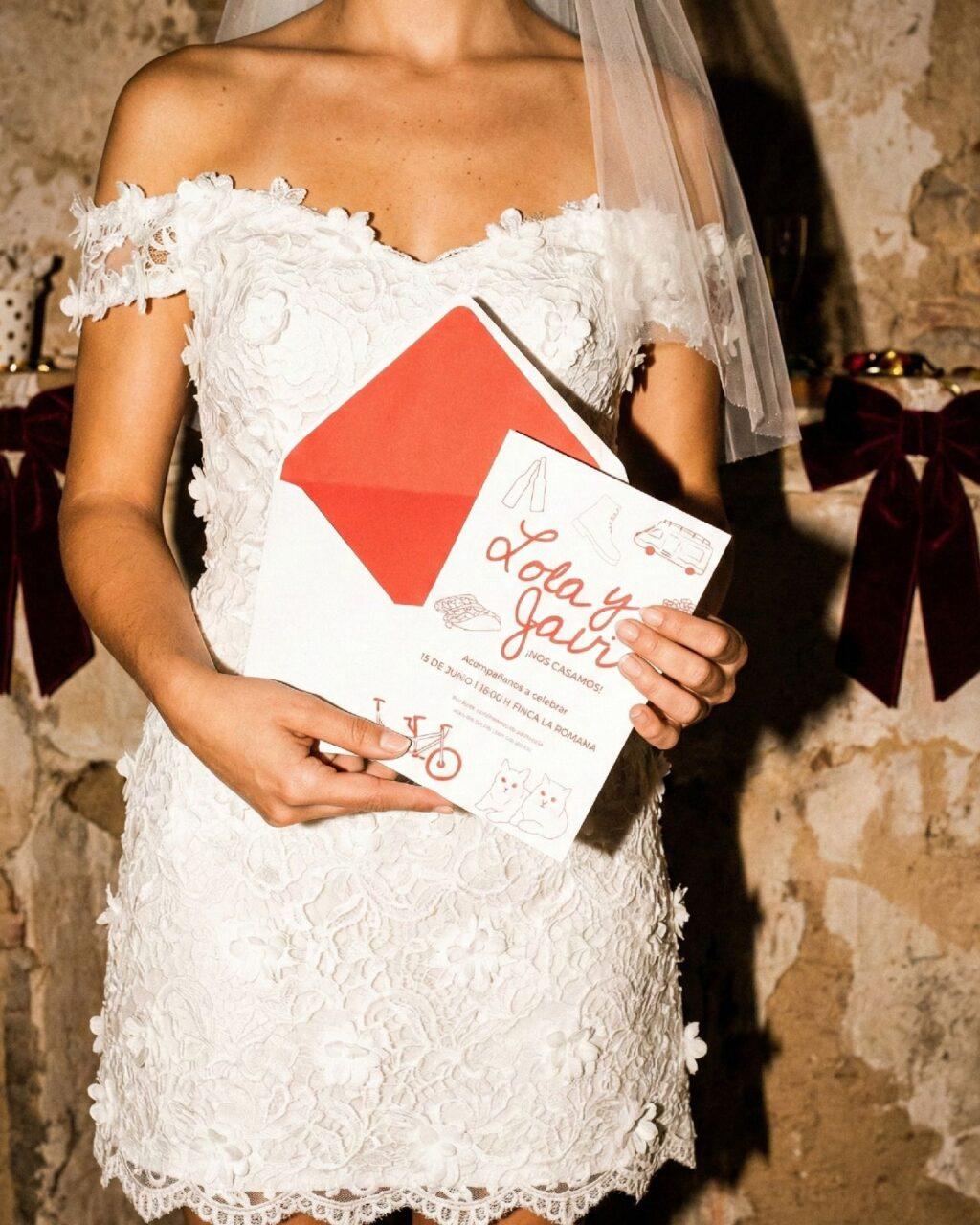 Novia con vestido bordado sosteniendo su invitación de boda Lola y Javi con sobre rojo