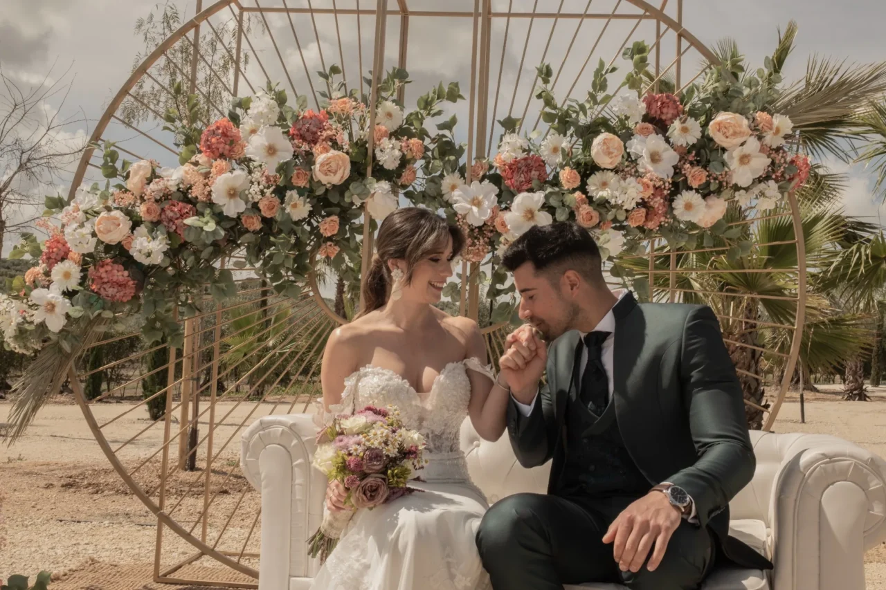 Novios sentados bajo un gran arco floral mientras el novio besa la mano de la novia en su boda